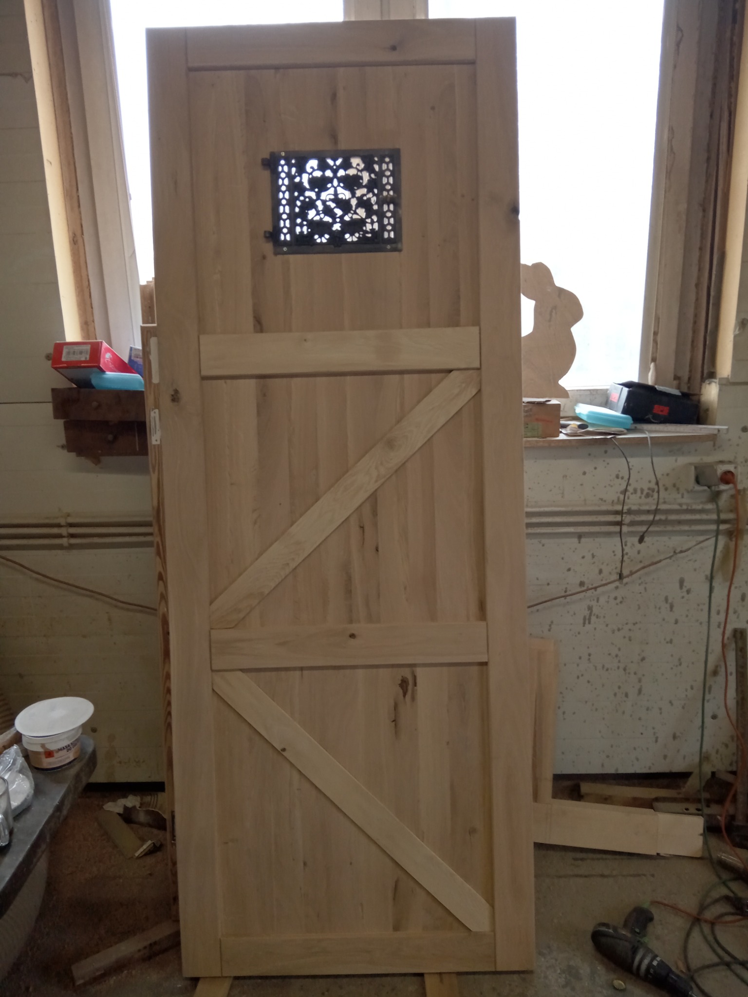 Surowa, niedokończona, drewniana brama z ozdobnym, metalowym okienkiem, oparta o ścianę w warsztacie.