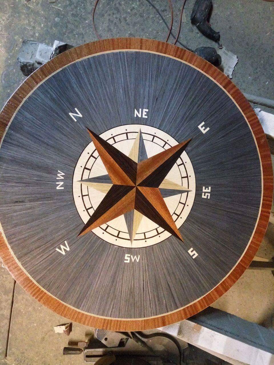 Okrągły blat stołu z intarsją w kształcie kompasu, wykonany z różnych gatunków drewna w odcieniach brązu i szarości, otoczony dekoracyjnym obrzeżem.