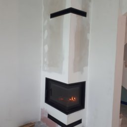 WIMAR Mariusz Jaremko - Wnętrze z narożnym wkładem kominkowym w trakcie montażu, widoczne niedoskonałości wykończenia ścian, ułożone drewno obok podstawy.