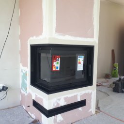 WIMAR Mariusz Jaremko - Wnętrze w trakcie wykańczania z wkładem kominkowym o czarnej ramie, widoczne naklejki informacyjne na szybie, ściany przygotowane pod malowanie, na podłodze folia ochronna i narzędzia.