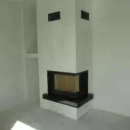 Nowoczesny narożny wkład kominkowy z czarnym obramowaniem, w surowym, białym wnętrzu z widocznymi śladami prac wykończeniowych na podłodze.