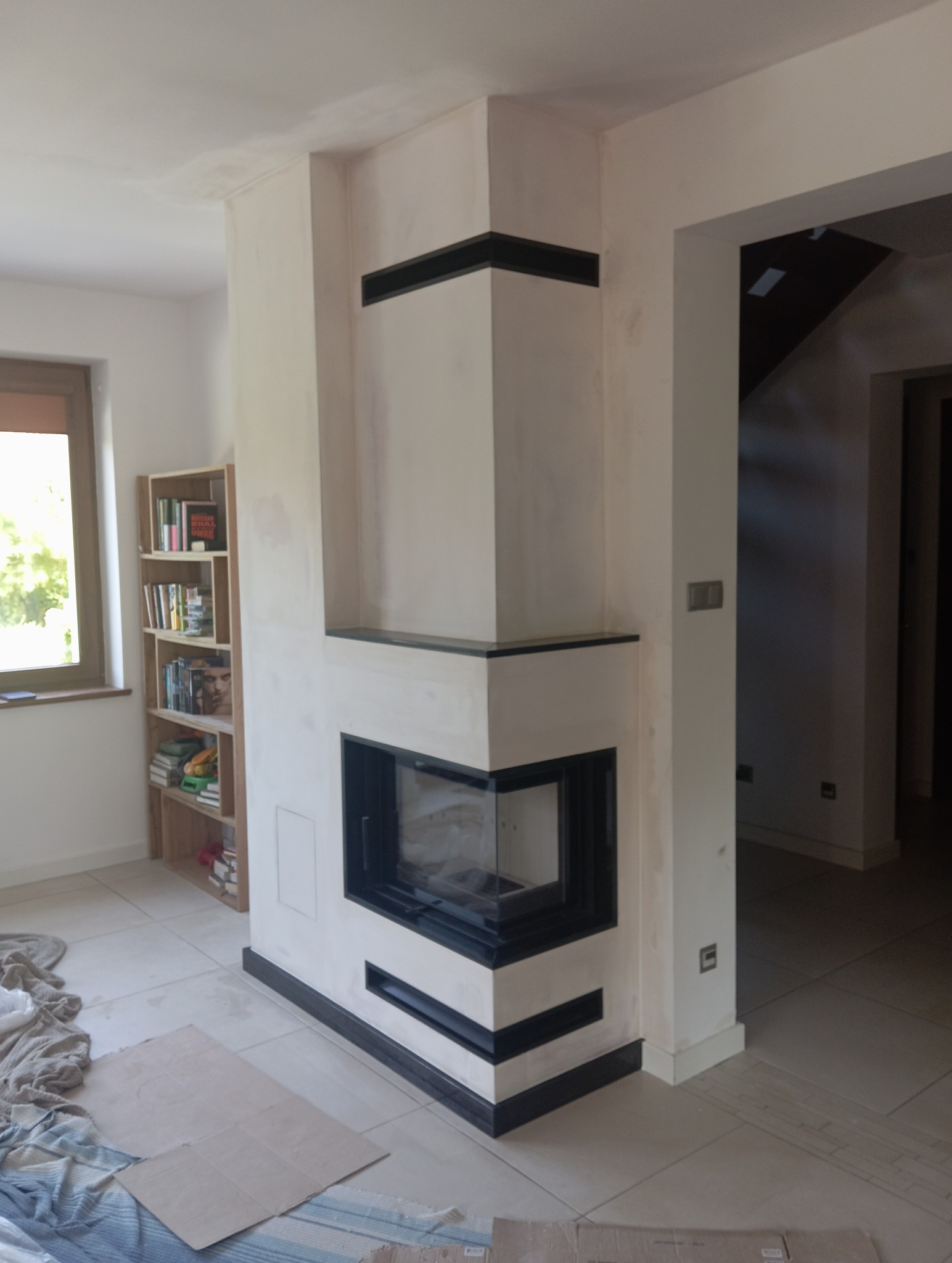 Narożny wkład kominkowy z czarnymi akcentami w trakcie zabudowy, widoczne niedoskonałości tynku i zabezpieczenia podłogi.