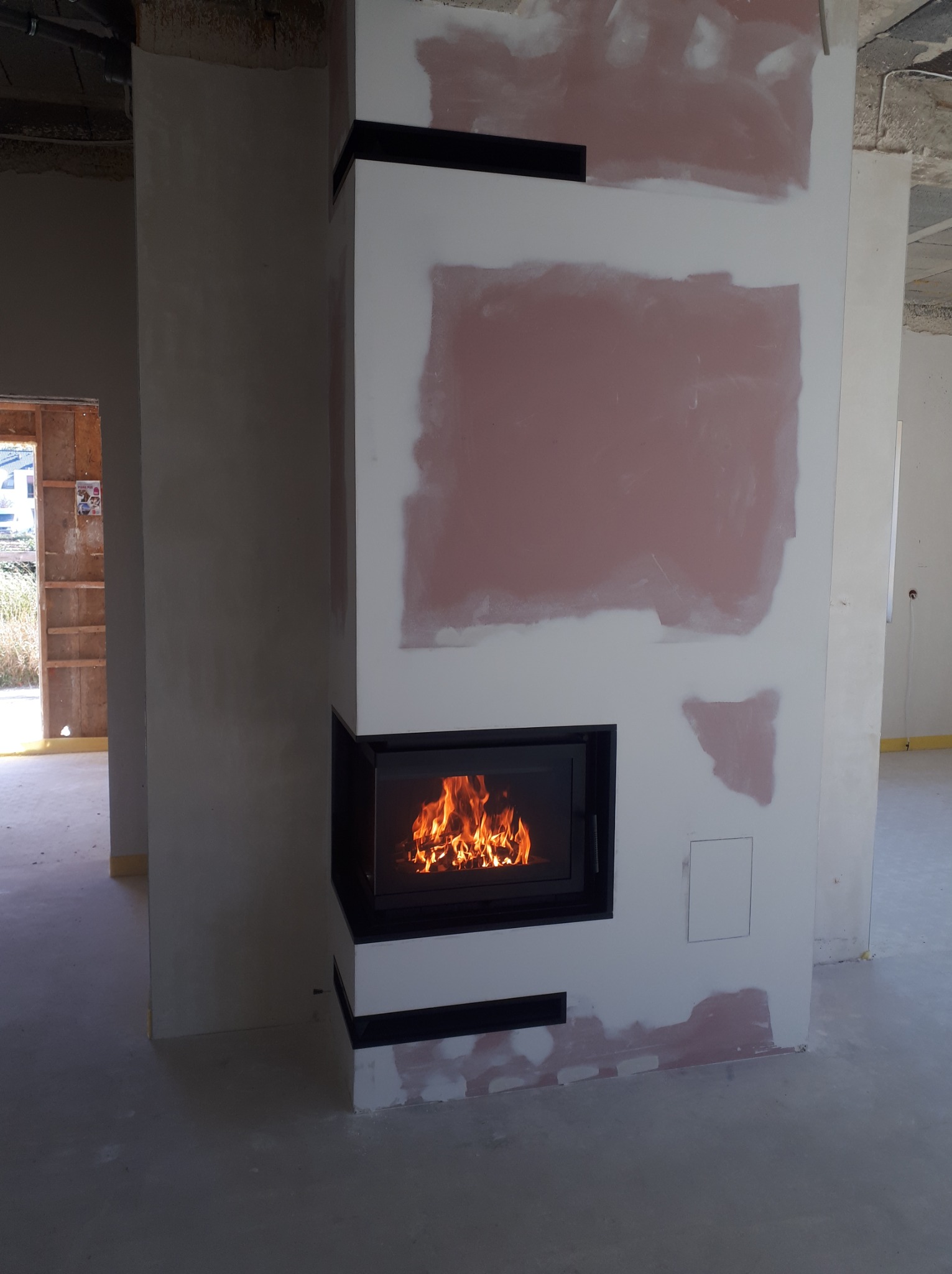 Wnętrze w trakcie wykańczania z narożnym wkładem kominkowym z czarną ramą, widoczne płomienie, ściany w stanie surowym, z widocznymi śladami szpachlowania.