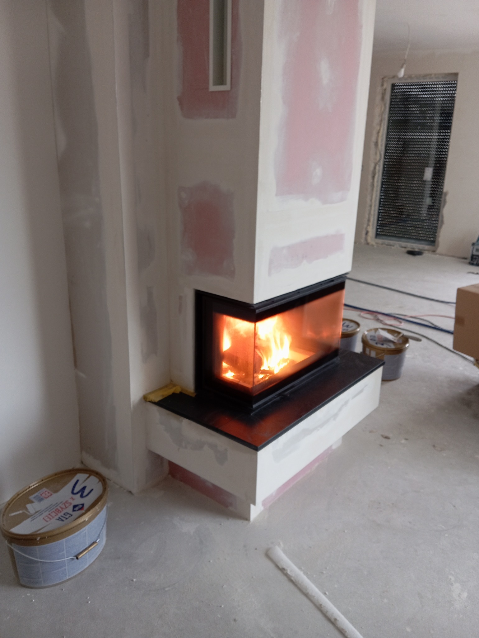 Niedokończona obudowa narożnego wkładu kominkowego z czarną ramą, widoczne płomienie, ślady szpachlowania na ścianach, puszka farby na podłodze, pomieszczenie w trakcie remontu.