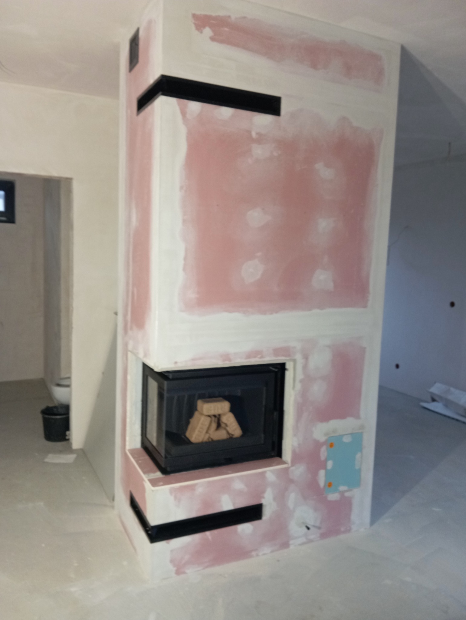 Wnętrze w trakcie wykańczania z widocznym obudowanym wkładem kominkowym, na ścianach ślady gładzi, czarne elementy dekoracyjne.