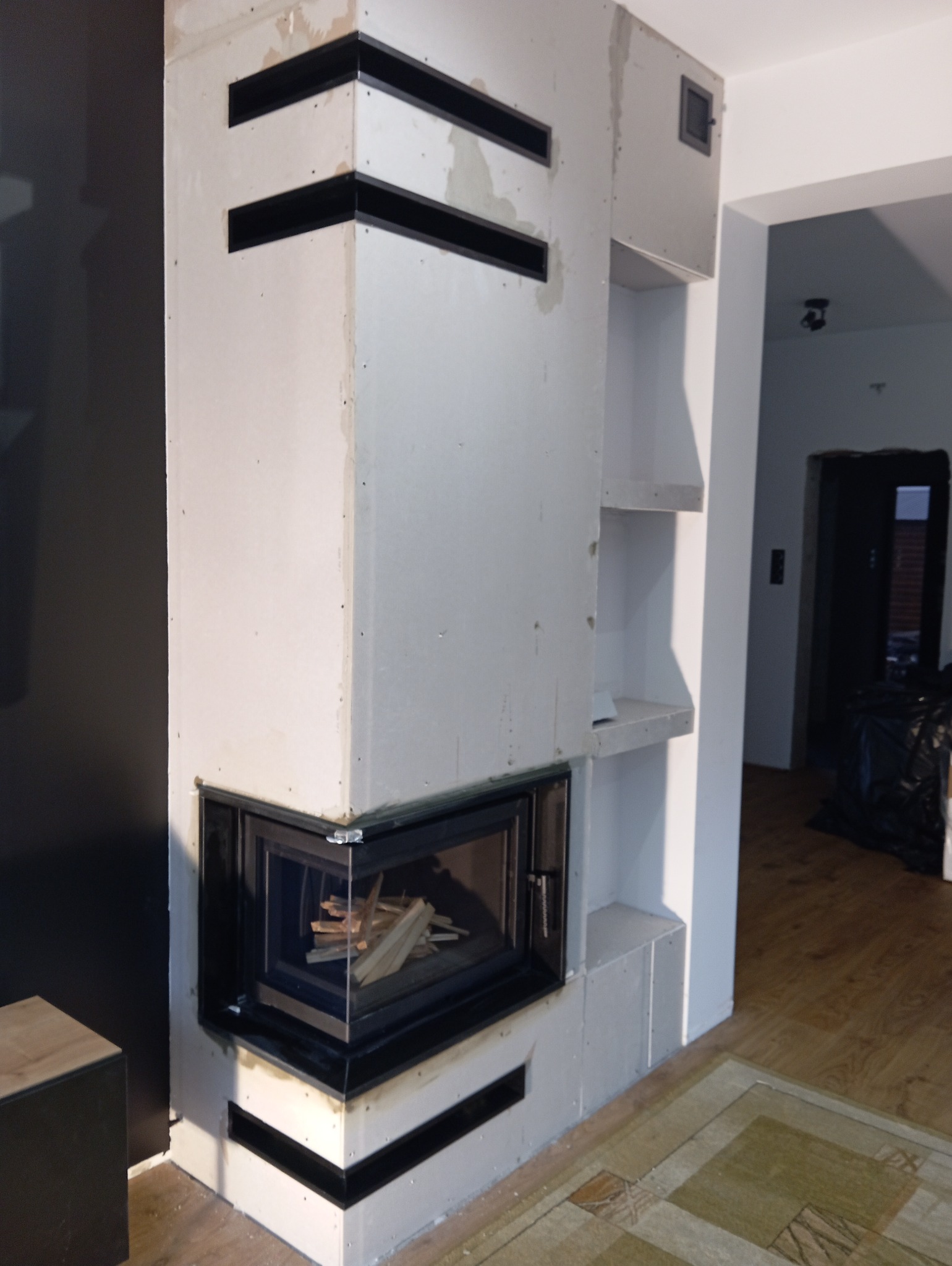 Niedokończona obudowa wnęki na palenisko z czarnymi akcentami, widoczne drewno w środku, surowe wykończenie gipsowe, półki w obudowie, widok na wnętrze domu z podłogą i dywanem.