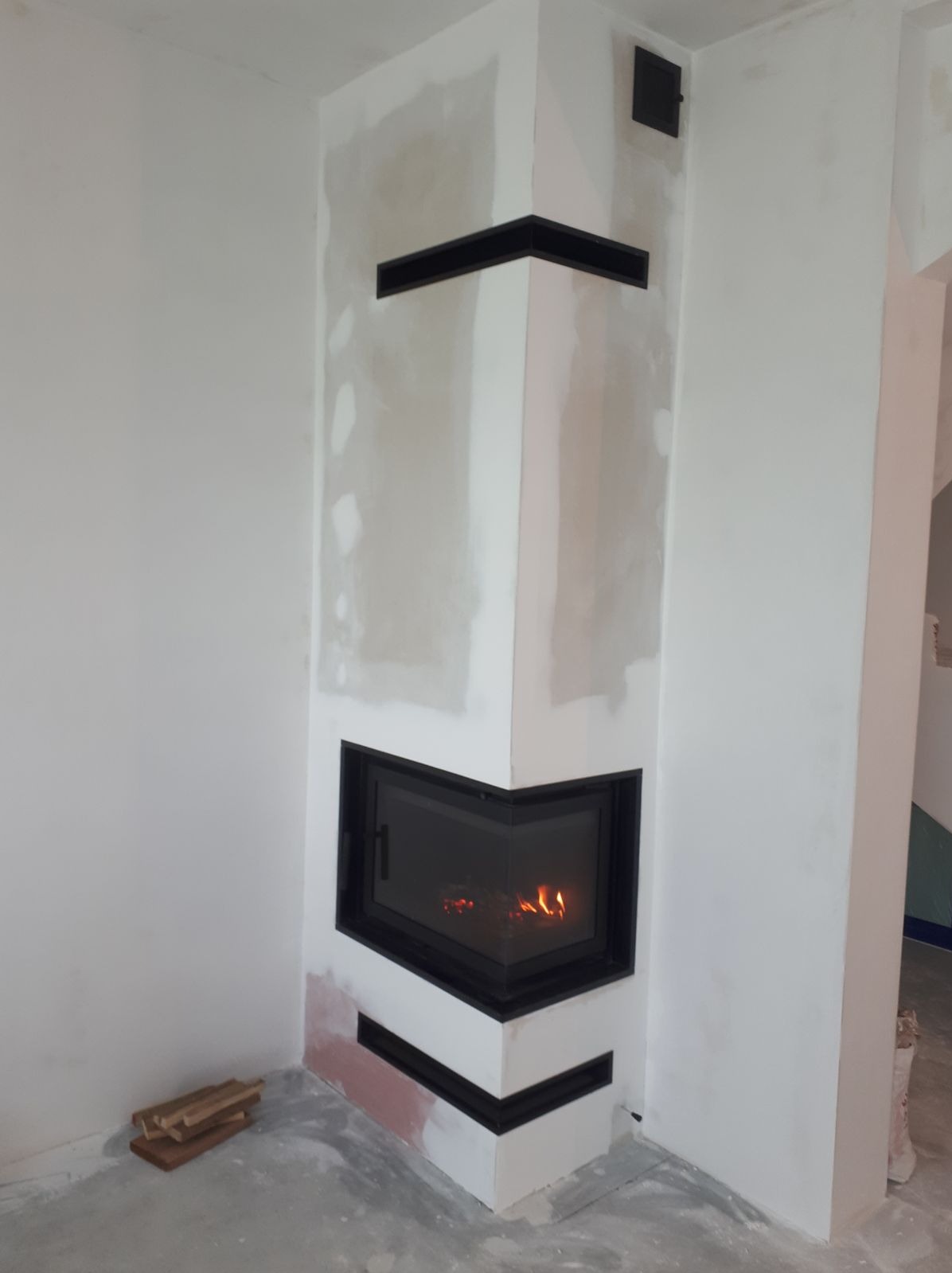 Wnętrze z narożnym wkładem kominkowym w trakcie montażu, widoczne niedoskonałości wykończenia ścian, ułożone drewno obok podstawy.