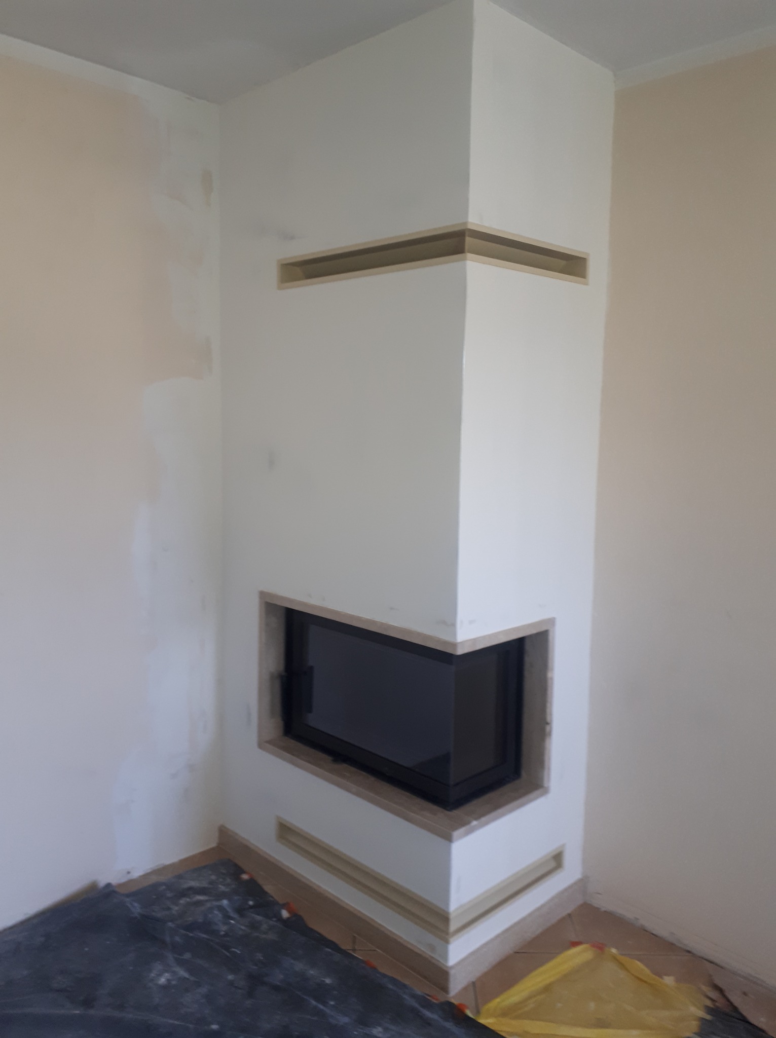 Narożny kominek w trakcie zabudowy, widoczne niedomalowane ściany i zabezpieczona podłoga.