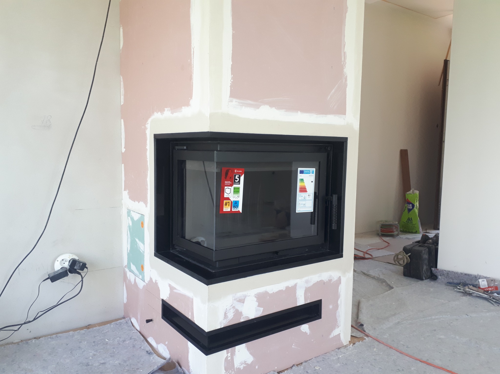 Wnętrze w trakcie wykańczania z wkładem kominkowym o czarnej ramie, widoczne naklejki informacyjne na szybie, ściany przygotowane pod malowanie, na podłodze folia ochronna i narzędzia.