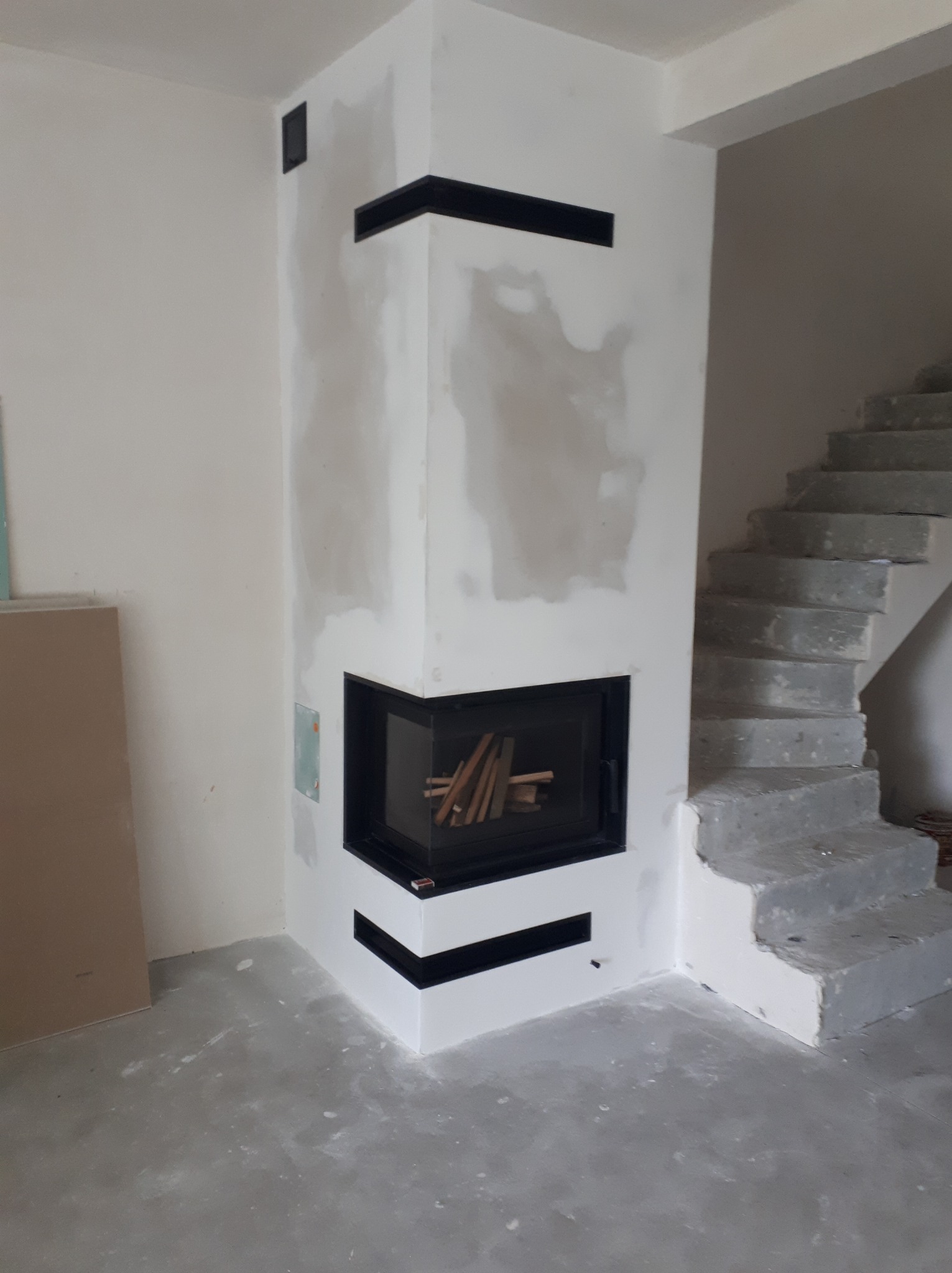Narożny wkład kominkowy z czarną ramą i dekoracyjnymi czarnymi pasami w obudowie, w trakcie wykańczania, widoczne surowe schody w tle.