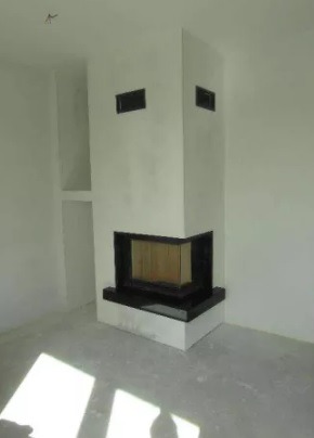 Nowoczesny narożny wkład kominkowy z czarnym obramowaniem, w surowym, białym wnętrzu z widocznymi śladami prac wykończeniowych na podłodze.