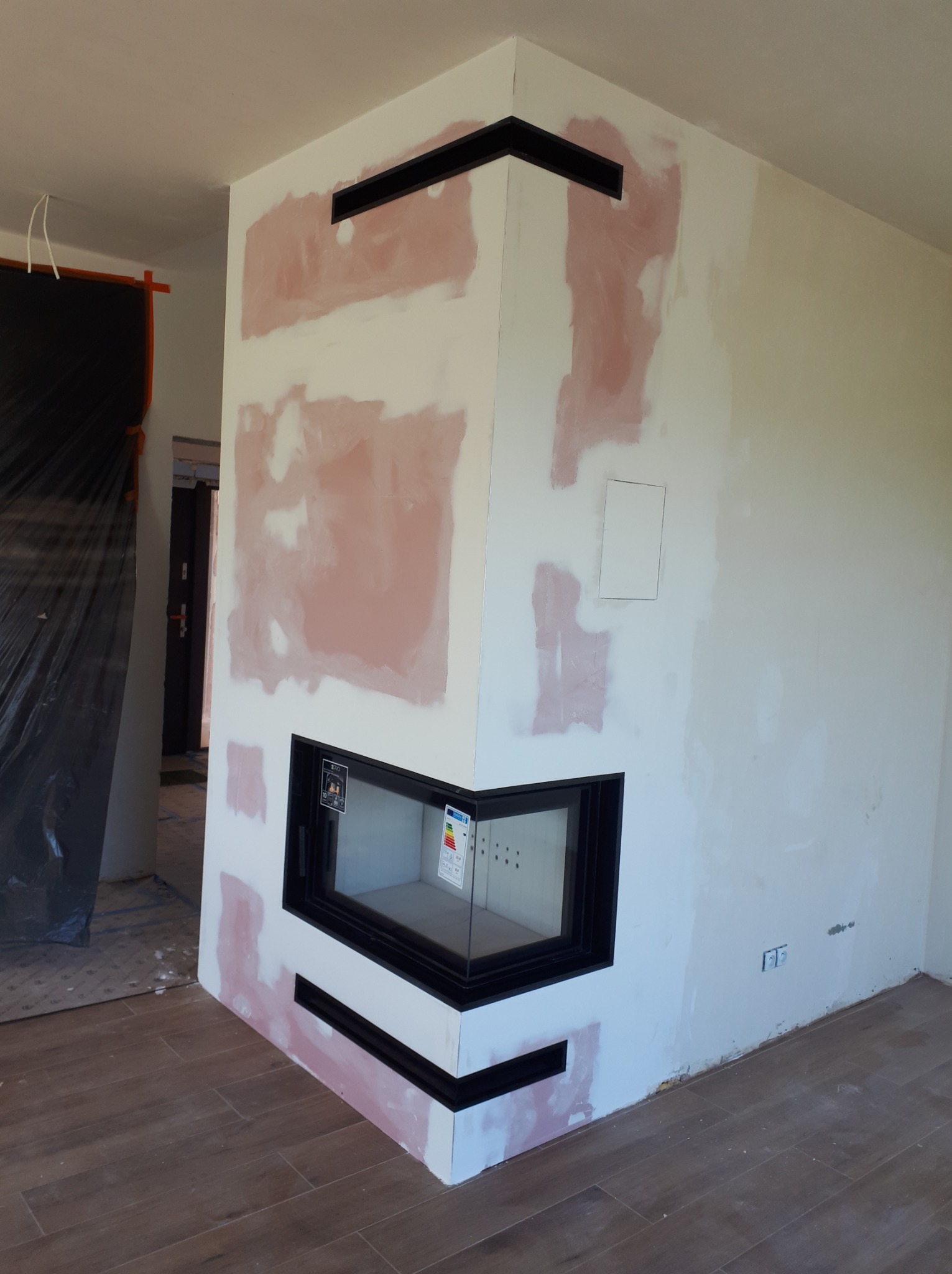 Wnętrze w trakcie wykończenia: obudowa wkładu kominkowego z czarną ramą i widocznymi śladami szpachlowania na ścianach. Widoczne elementy instalacji elektrycznej i fragment podłogi z paneli.