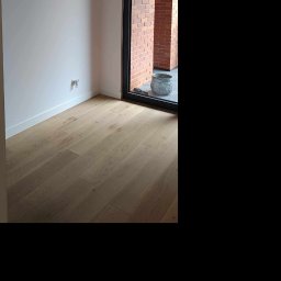 Renova - Jasne panele podłogowe w pokoju po remoncie w Warszawie. Widoczne białe ściany i okno z widokiem na ceglany mur. Dekoracyjna donica przy oknie.