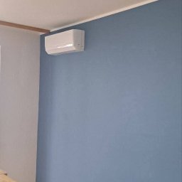 Renova - Błękitna ściana po odświeżeniu z zamontowanym klimatyzatorem, fragment drewnianej komody i podłogi. Widoczny kontakt elektryczny.