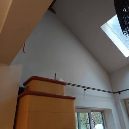 Renova - Jasne wnętrze z ukośnym sufitem i oknem dachowym. Widoczny piec kaflowy w odcieniach żółci i brązu, oraz karnisz na tle białej ściany i okna.