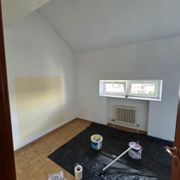 Renova - Pokój w trakcie malowania: ściana z fragmentem pomalowanym na beżowo, zabezpieczona podłoga, farby, wałek. Widok na okno i grzejnik, sufit ze skosem.