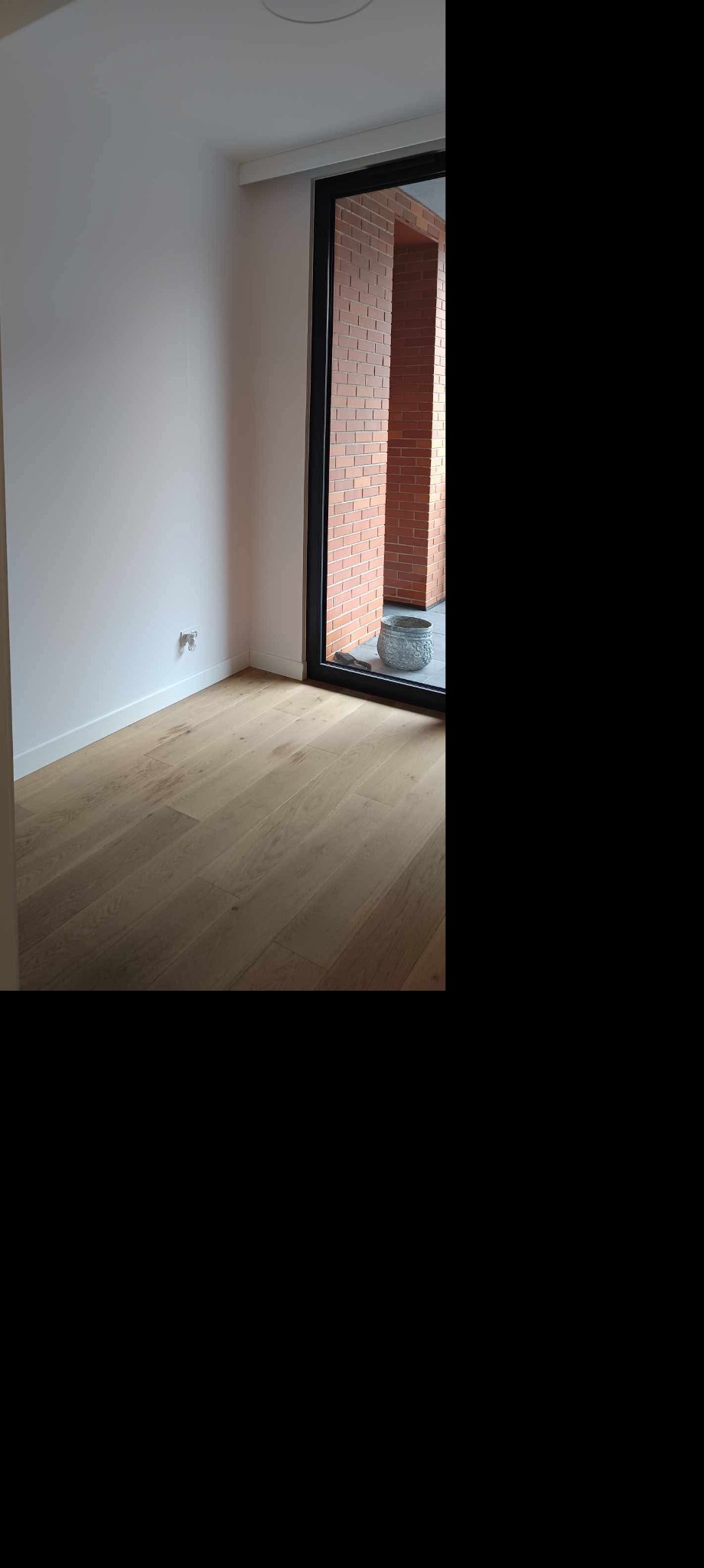 Jasne panele podłogowe w pokoju po remoncie w Warszawie. Widoczne białe ściany i okno z widokiem na ceglany mur. Dekoracyjna donica przy oknie.