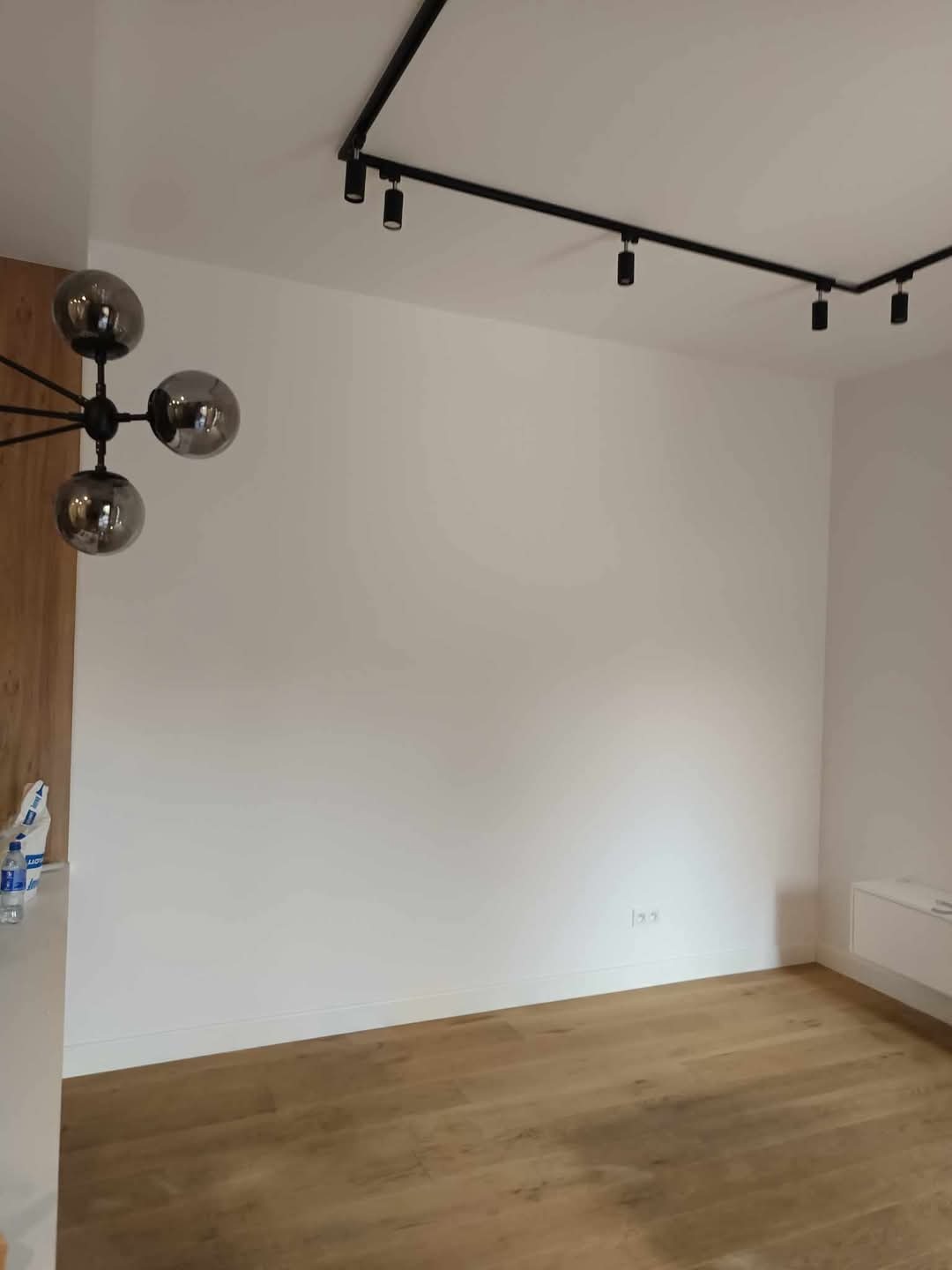 Jasne wnętrze po odświeżeniu: białe ściany, drewniana podłoga, nowoczesne oświetlenie szynowe. W rogu widoczny fragment lampy.