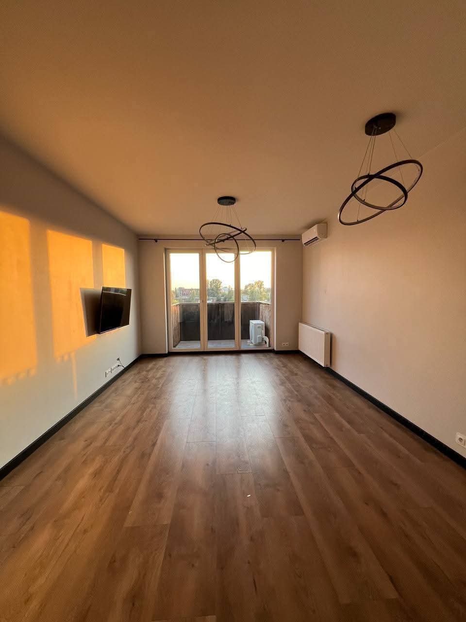 Jasne, puste wnętrze z drewnianą podłogą, nowoczesnymi lampami sufitowymi i balkonem z widokiem na zieleń. Ściana po lewej oświetlona zachodzącym słońcem.