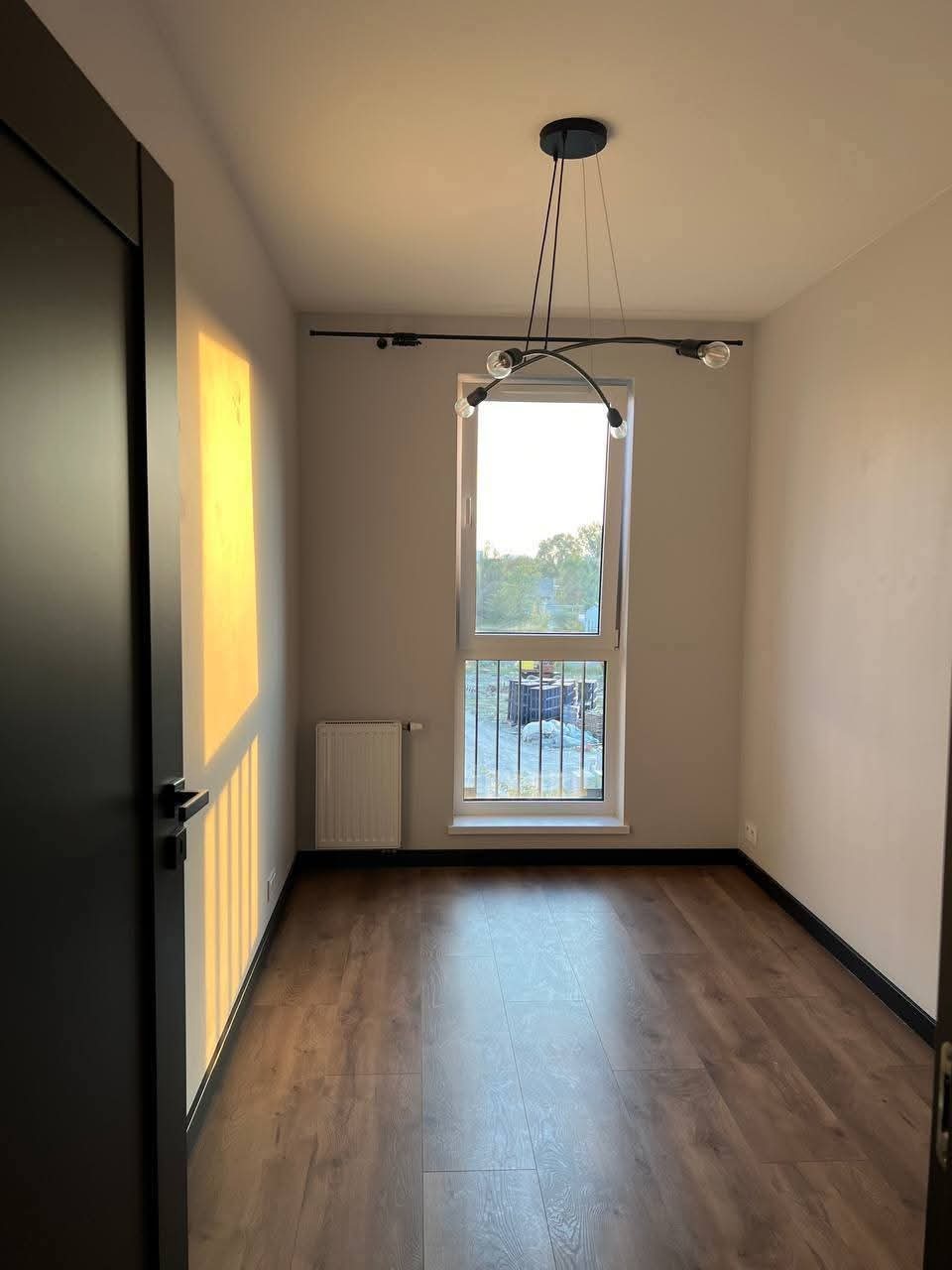 Puste wnętrze pokoju z drewnianą podłogą, oświetlone przez okno z widokiem na plac budowy. Nowoczesna lampa sufitowa. Minimalistyczny wystrój.