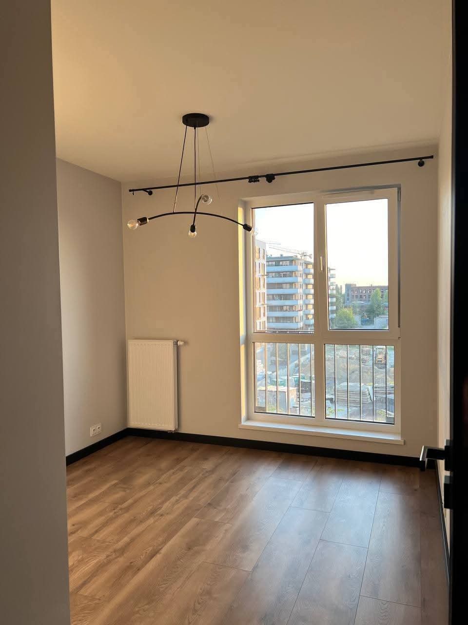 Jasne wnętrze z panelami podłogowymi, grzejnikiem i oknem z widokiem na miasto. Minimalistyczna lampa sufitowa dodaje nowoczesnego charakteru.