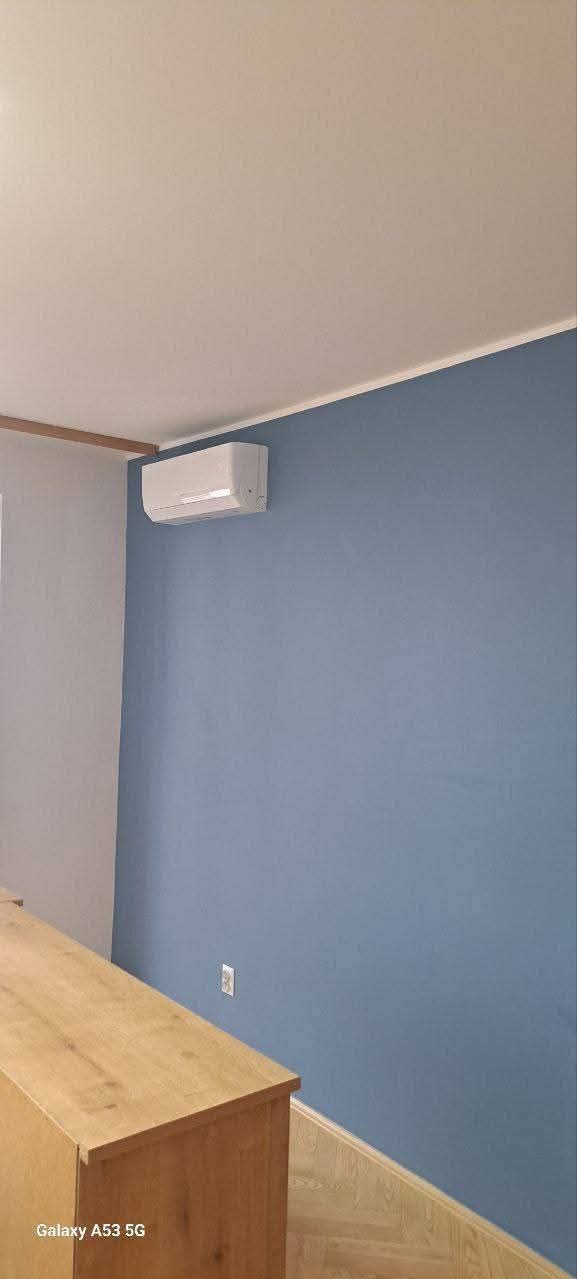 Błękitna ściana po odświeżeniu z zamontowanym klimatyzatorem, fragment drewnianej komody i podłogi. Widoczny kontakt elektryczny.