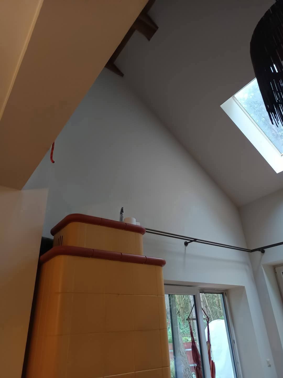 Jasne wnętrze z ukośnym sufitem i oknem dachowym. Widoczny piec kaflowy w odcieniach żółci i brązu, oraz karnisz na tle białej ściany i okna.