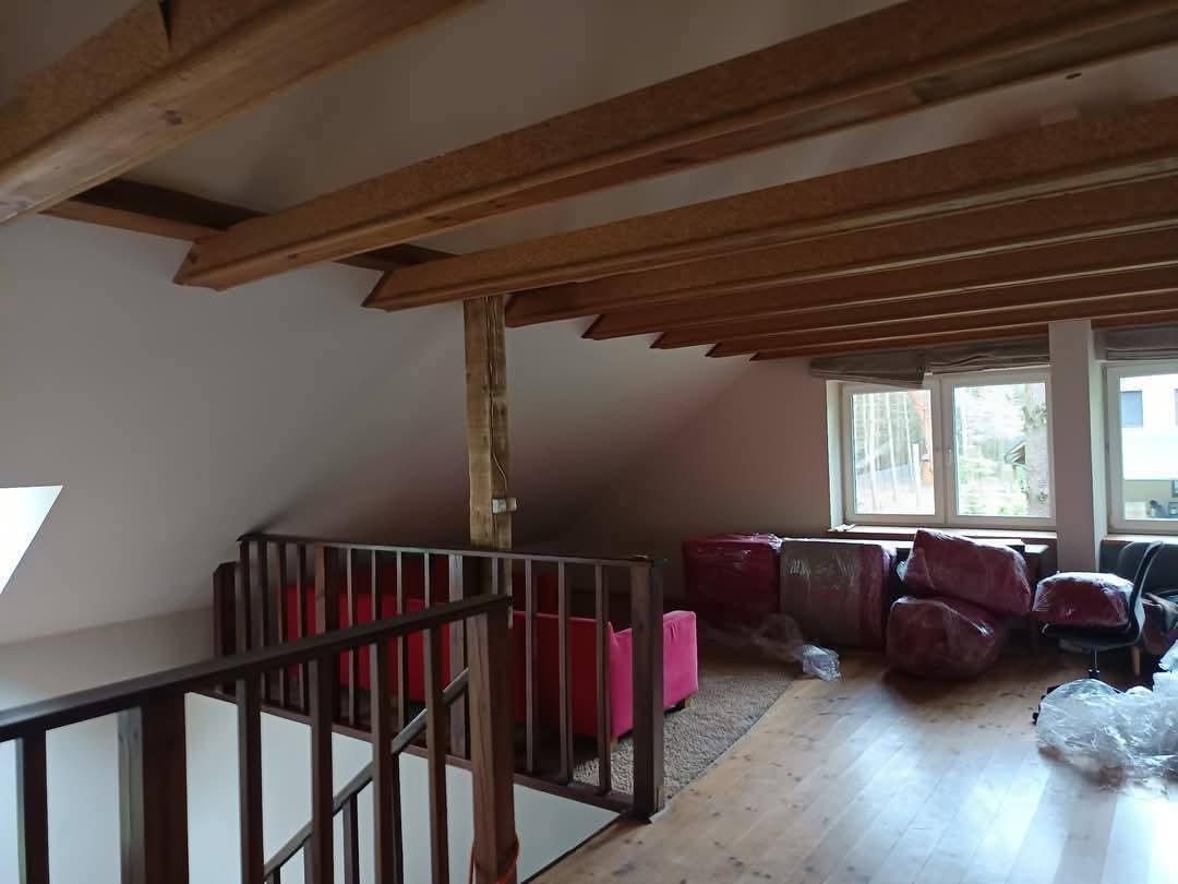 Poddasze z drewnianą konstrukcją stropu w trakcie przygotowań do prac wykończeniowych. Meble zabezpieczone folią, widoczne okna i barierka schodów.