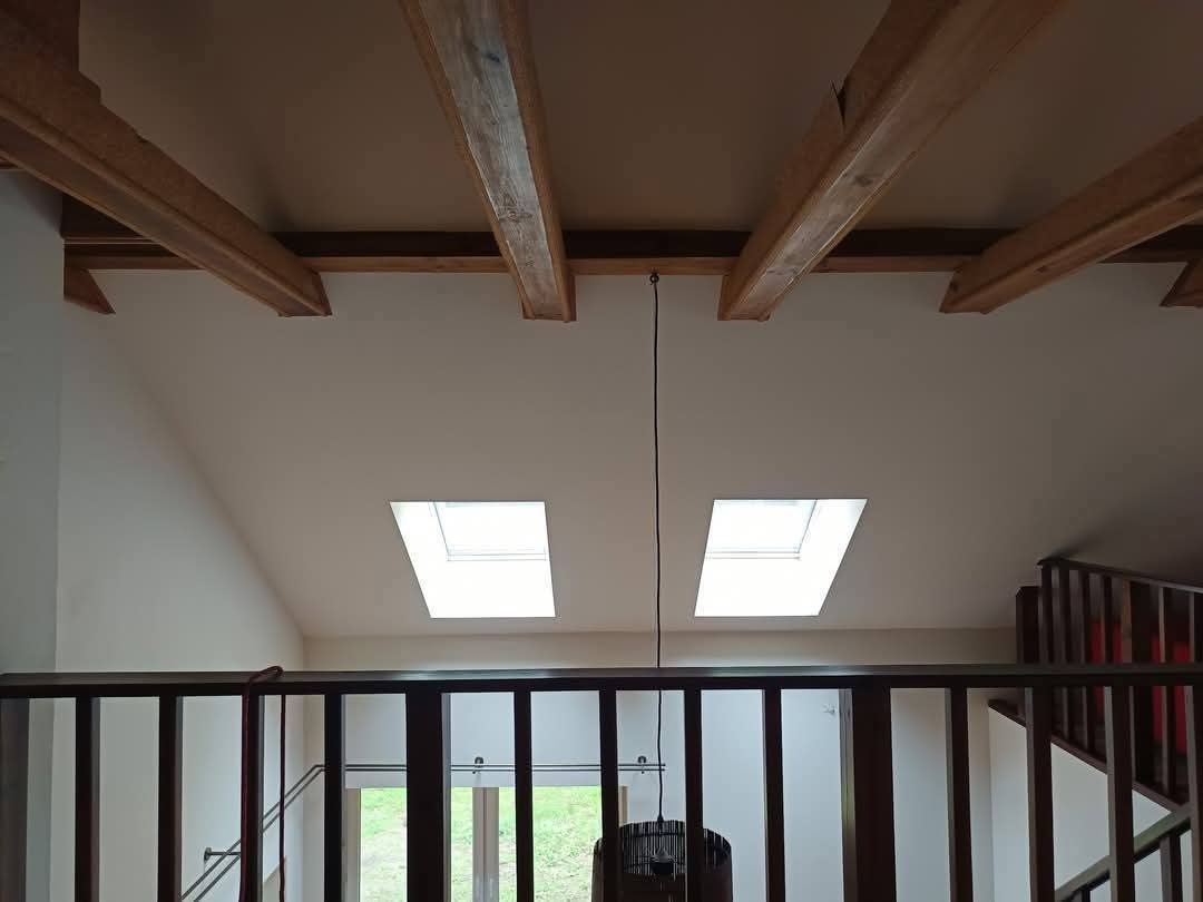 Jasne poddasze z drewnianymi belkami i dwoma oknami dachowymi, widoczne zza balustrady. Ściany w odcieniach bieli, schody z drewnianą barierką po prawej.