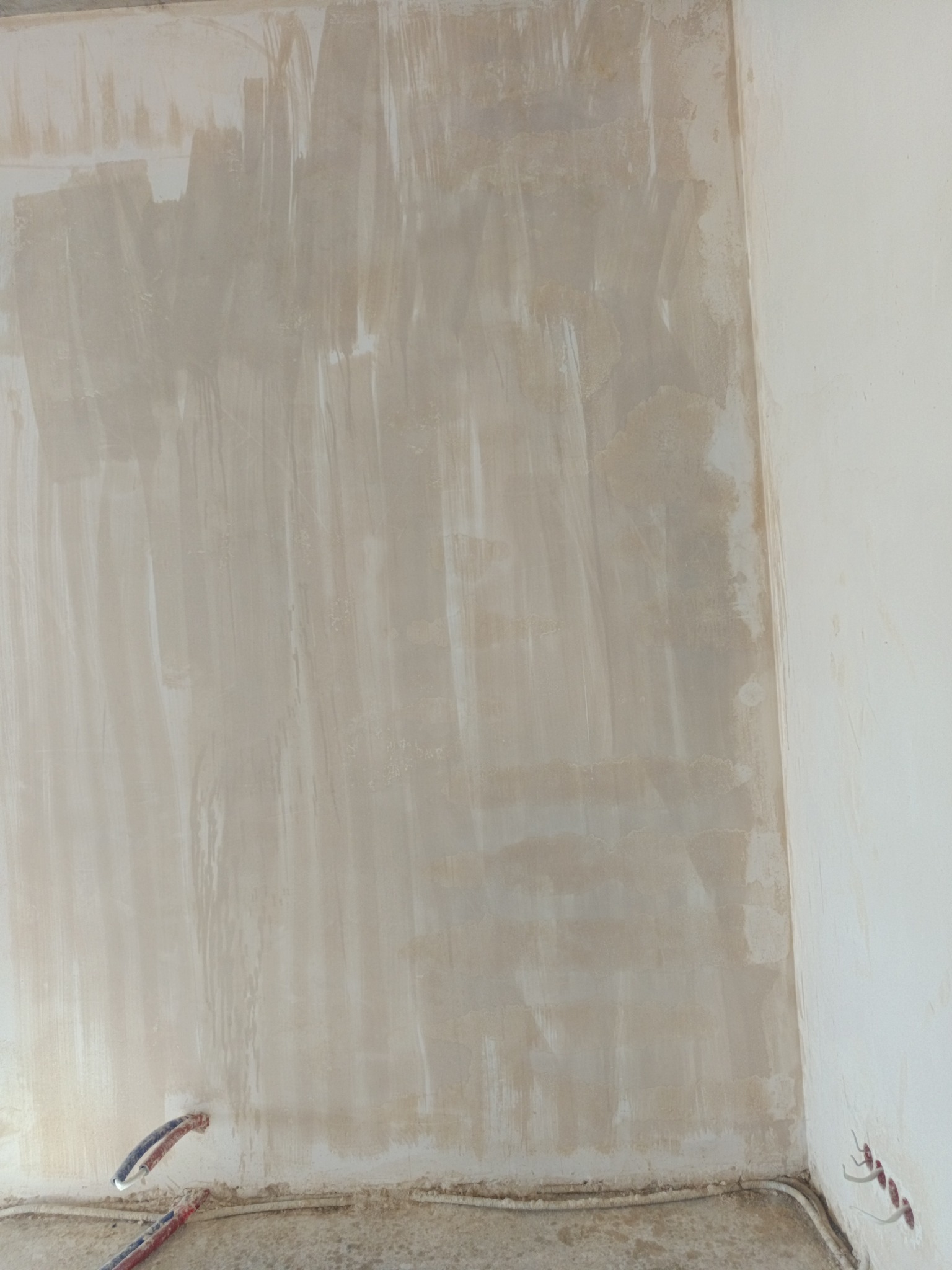 Ściana po nałożeniu pierwszej warstwy gładzi szpachlowej, widoczne przewody elektryczne przy podłodze. Nierównomierne krycie, przygotowanie do dalszych prac.