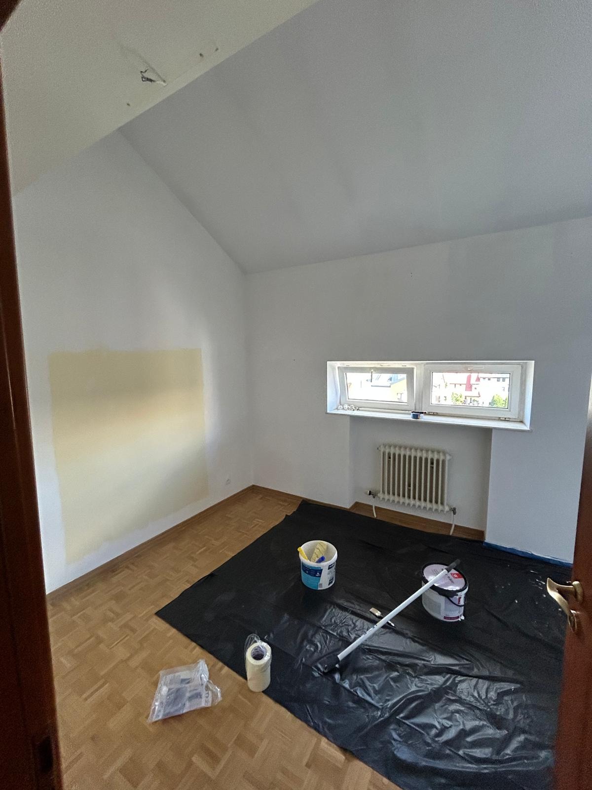 Pokój w trakcie malowania: ściana z fragmentem pomalowanym na beżowo, zabezpieczona podłoga, farby, wałek. Widok na okno i grzejnik, sufit ze skosem.