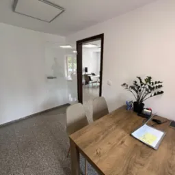 Jasne wnętrze biura z drewnianym biurkiem, rośliną doniczkową i dokumentami, widok na przeszkloną przestrzeń biurową z pracownikiem w tle.