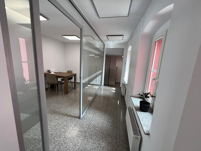 Jasne wnętrze biurowe z przeszklonymi ścianami, widok na salę konferencyjną z drewnianym stołem i krzesłami, korytarz z oknami w kształcie łuków i rośliną doniczkową na parapecie.