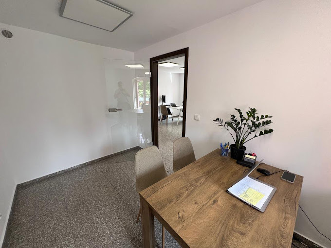 Jasne wnętrze biura z drewnianym biurkiem, rośliną doniczkową i dokumentami, widok na przeszkloną przestrzeń biurową z pracownikiem w tle.