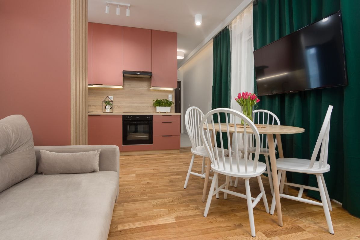 Jasne wnętrze mieszkania z różową zabudową kuchenną, okrągłym stołem z białymi krzesłami, szarą sofą i zielonymi zasłonami, oświetlone trzema punktowymi lampami sufitowymi.