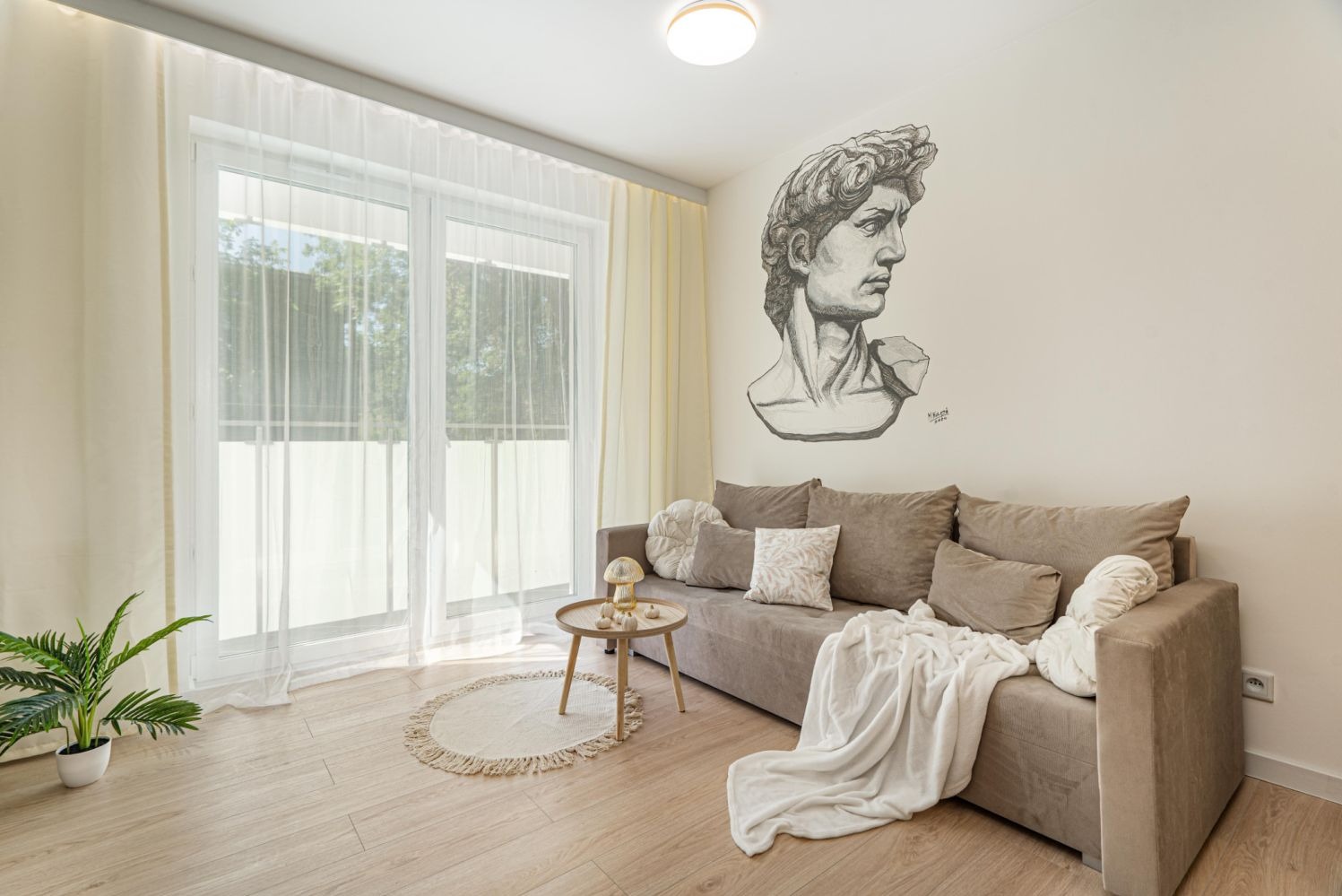 Jasny salon z beżową sofą, okrągłym dywanikiem, rośliną w doniczce i rysunkiem głowy Dawida na ścianie