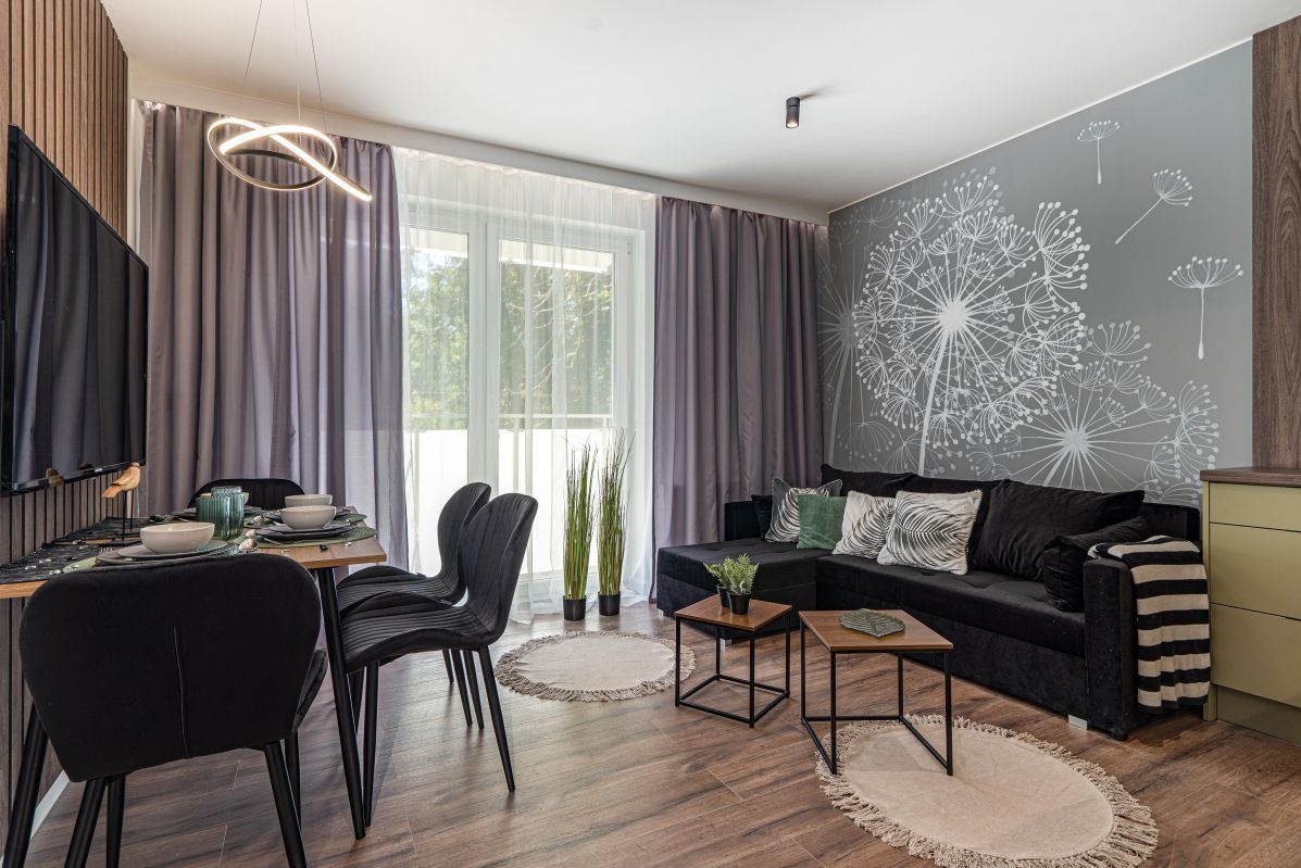 Nowoczesny salon z czarną sofą narożną, szarymi zasłonami i tapetą w dmuchawce; stół z nakryciem i czarnymi krzesłami, drewniana podłoga.