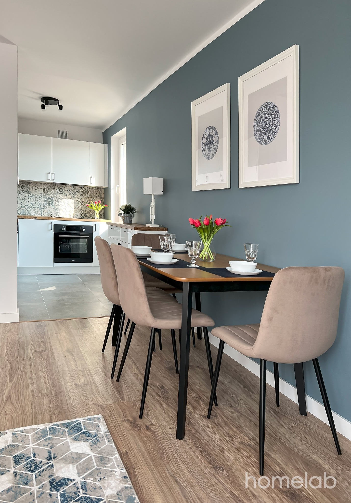 Nowoczesna jadalnia z drewnianym stołem nakrytym dla czterech osób, z bukietem czerwonych tulipanów w wazonie, beżowymi krzesłami na czarnych nogach, szarą ścianą z dwoma obrazami w białych ramach,...