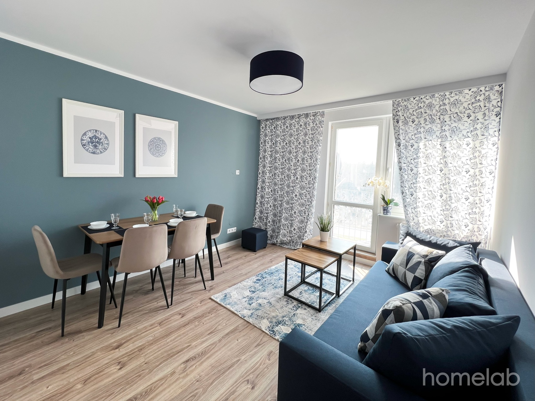 Jasne wnętrze mieszkania z drewnianą podłogą, niebieską sofą z wzorzystymi poduszkami, stołem z krzesłami i dekoracjami na ścianie, oświetlone naturalnym światłem wpadającym przez okno...