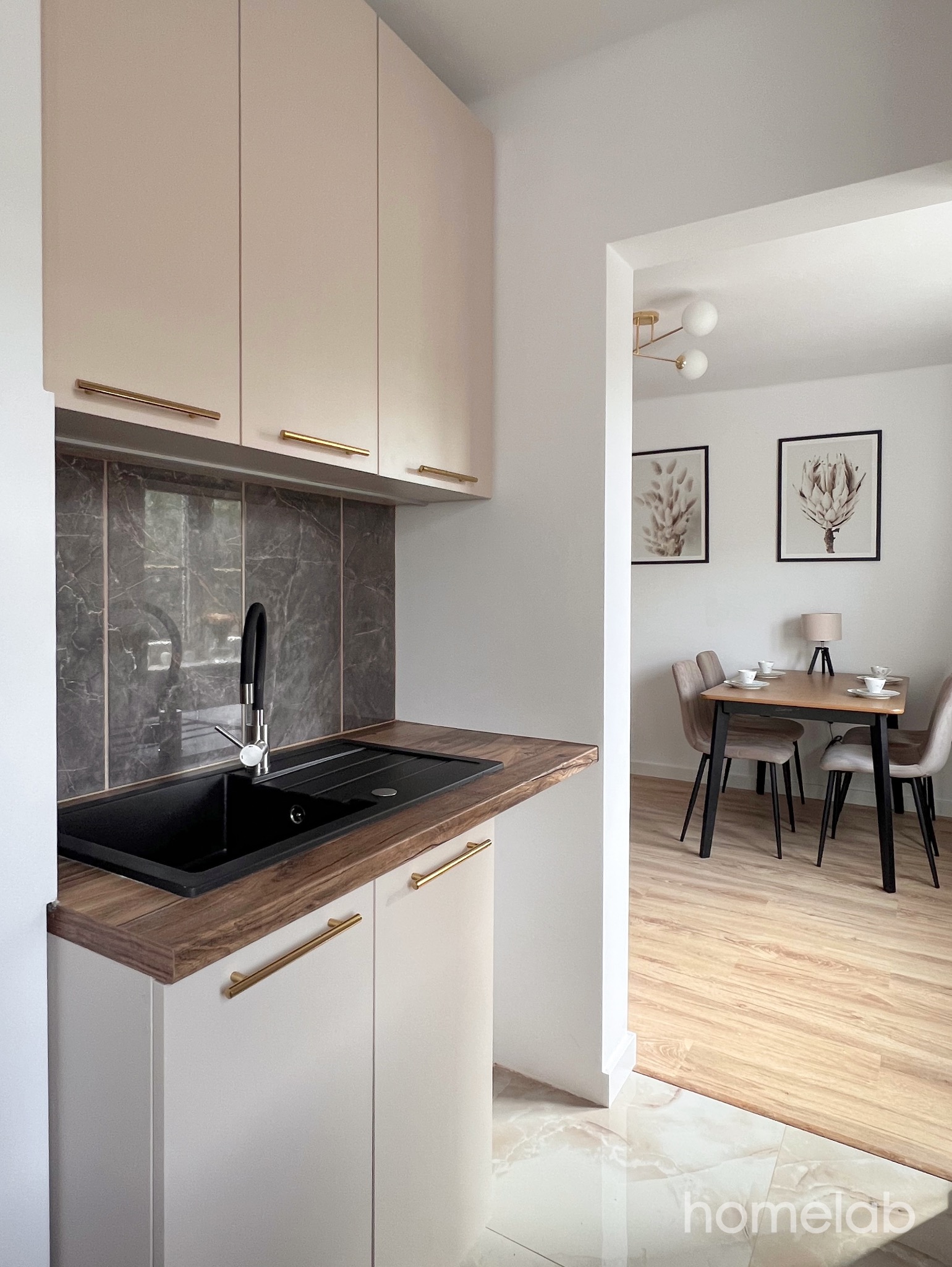 Nowoczesna kuchnia z beżowymi szafkami, czarnym zlewem i blatem imitującym drewno, widok na jadalnię z drewnianym stołem i beżowymi krzesłami, uzupełniona obrazami na ścianie.