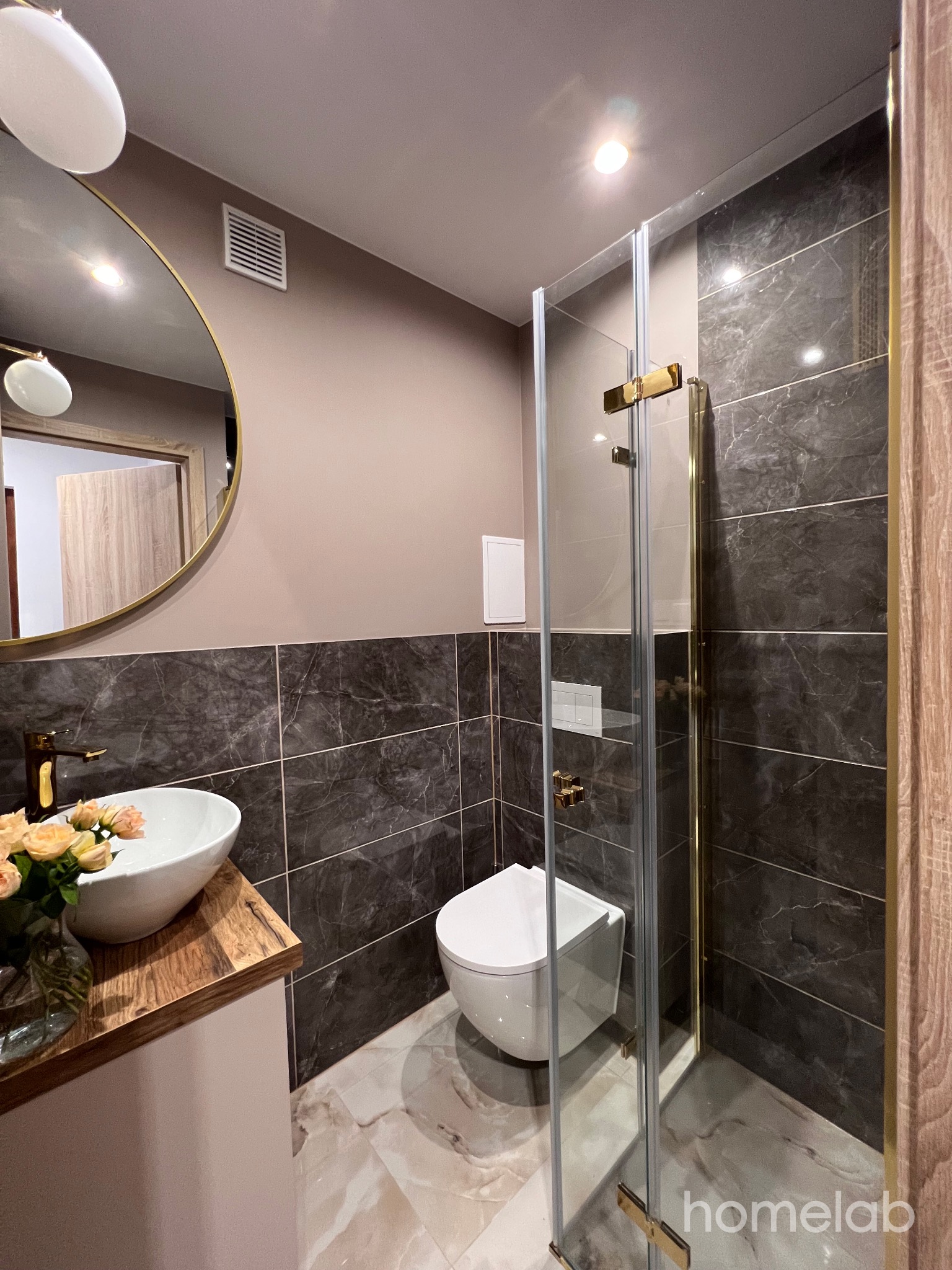 Nowoczesna łazienka z marmurowymi płytkami, okrągłym lustrem z złotą ramą, szklaną kabiną prysznicową z złotymi zawiasami i umywalką nablatową z bukietem różowych róż.