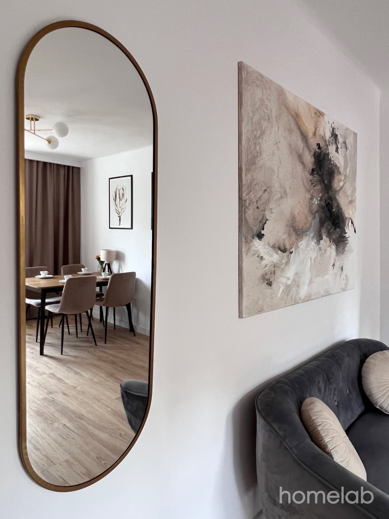Salon z szarym narożnikiem, abstrakcyjnym obrazem i lustrem w złotej ramie odbijającym jadalnię z drewnianym stołem i krzesłami w odcieniach beżu.