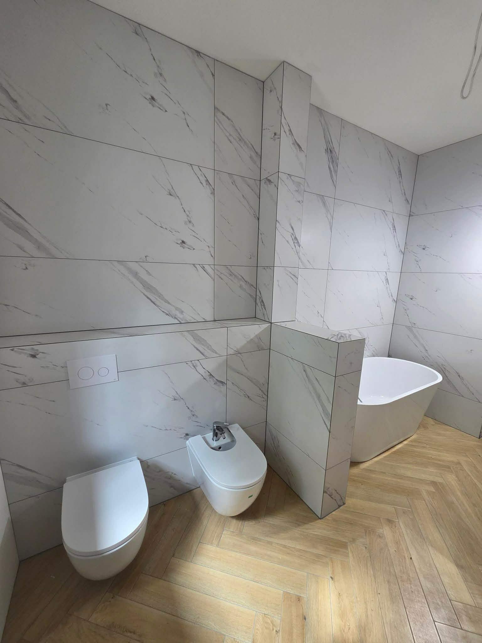 Nowoczesna łazienka z białymi płytkami imitującymi marmur, drewnianą podłogą w jodełkę, wiszącą toaletą i bidetem oraz wolnostojącą wanną.