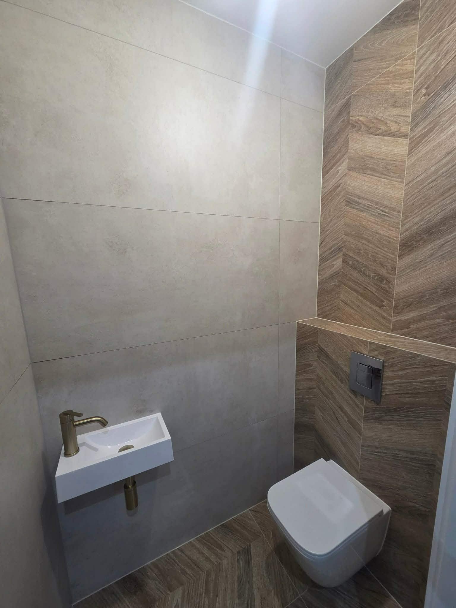 Nowoczesna łazienka z beżowymi płytkami na ścianach i podłodze ułożonymi w jodełkę. Umywalka narożna i podwieszana toaleta.