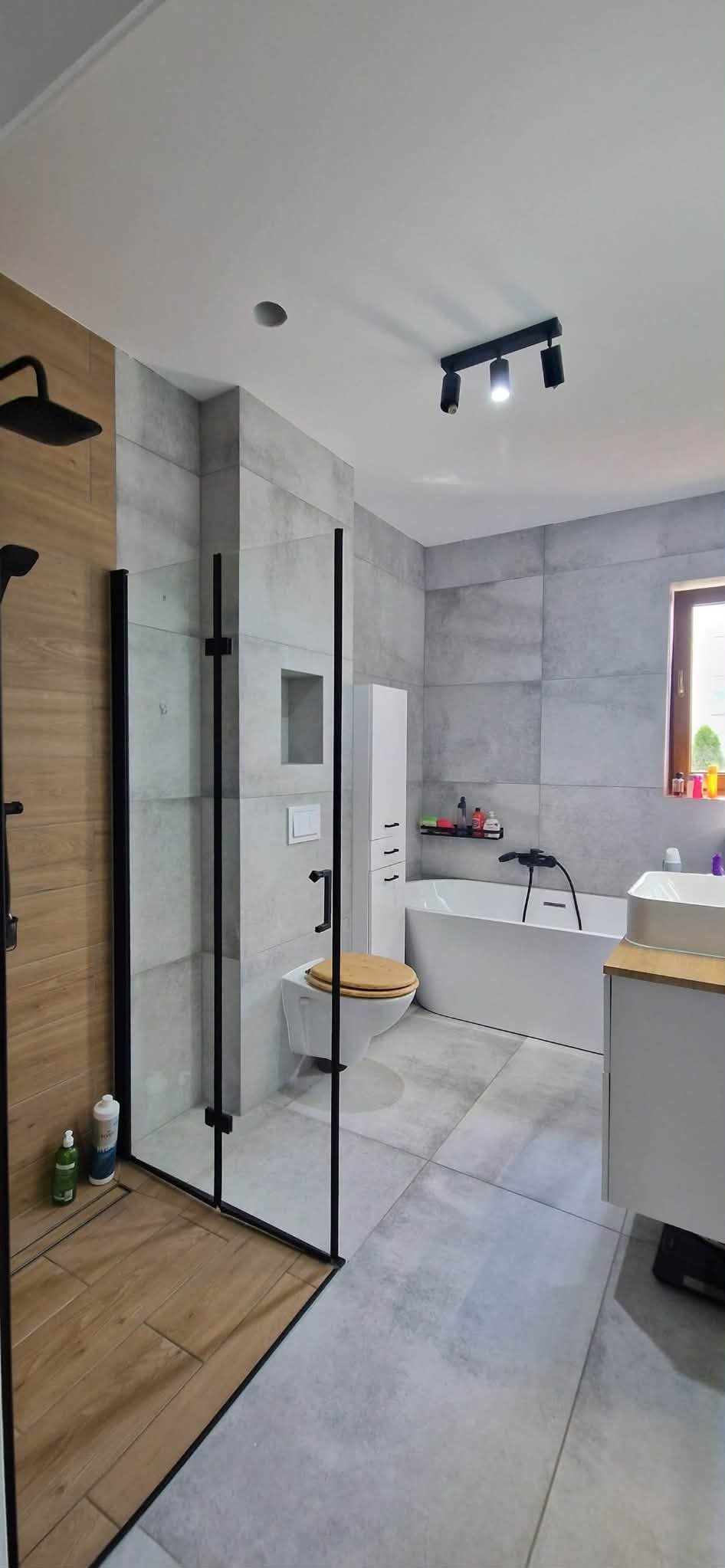 Nowoczesna łazienka z kabiną prysznicową typu walk-in, szare płytki wielkoformatowe, drewnopodobne akcenty i minimalistyczne oświetlenie punktowe.