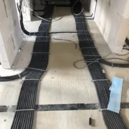 Równolegle ułożone wiązki peszli z kablami elektrycznymi, przymocowane metalowymi opaskami do surowej posadzki w trakcie instalacji w nowym budynku.