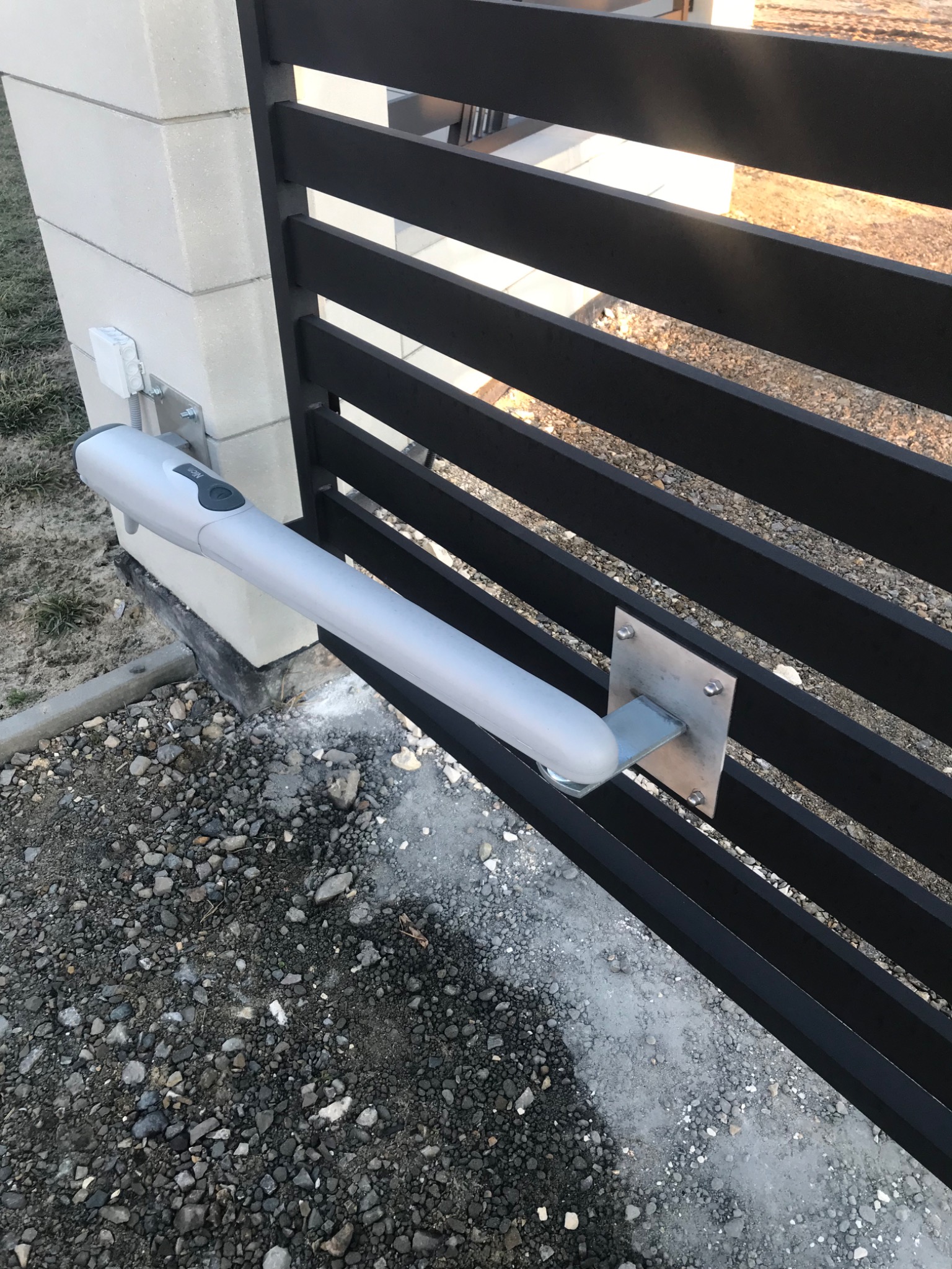 Szary siłownik automatycznej bramy przymocowany do czarnego, metalowego ogrodzenia na tle betonowego słupka i żwirowego podłoża.