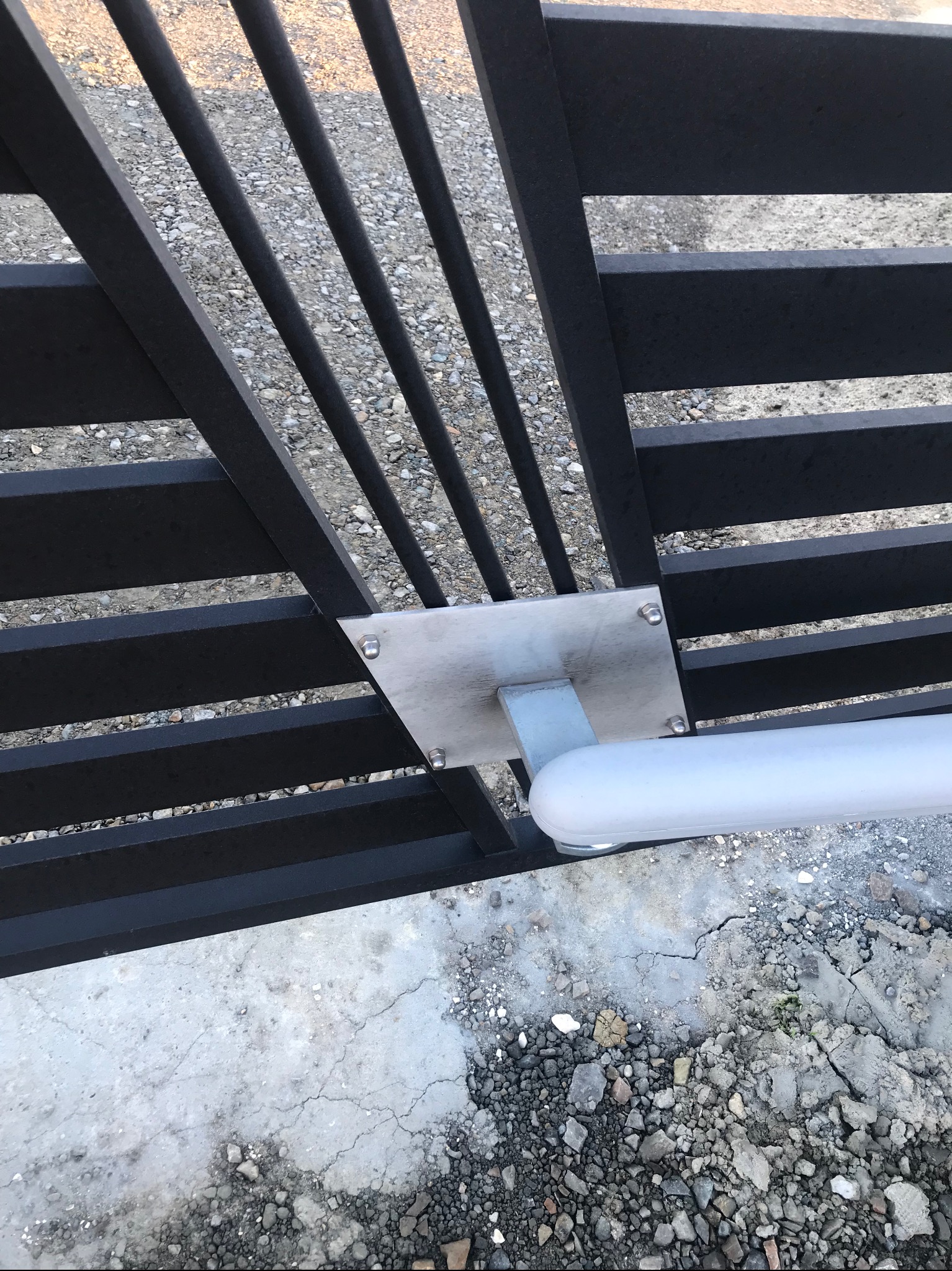 Fragment czarnej, metalowej bramy z automatyką, widoczny mechanizm napędu z osprzętem montażowym na tle betonowej nawierzchni z drobnymi kamieniami.