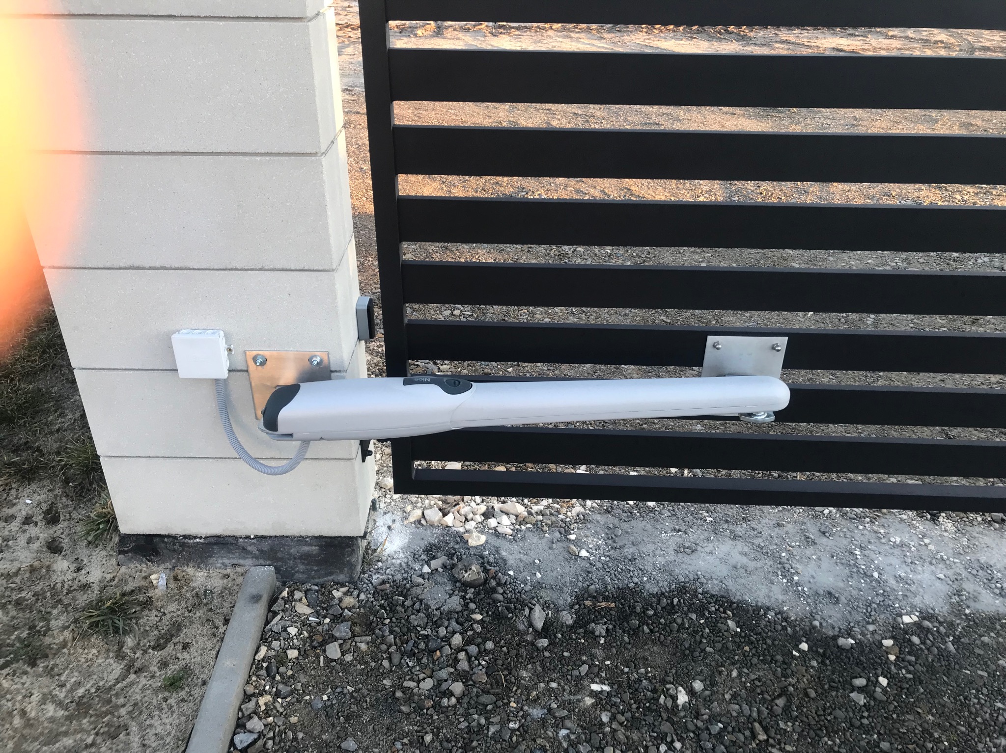 Automatyka bramy przesuwnej NICE zamontowana na słupku z bloczków betonowych i czarnej, poziomej bramie, widoczne przewody elektryczne w białej puszce.