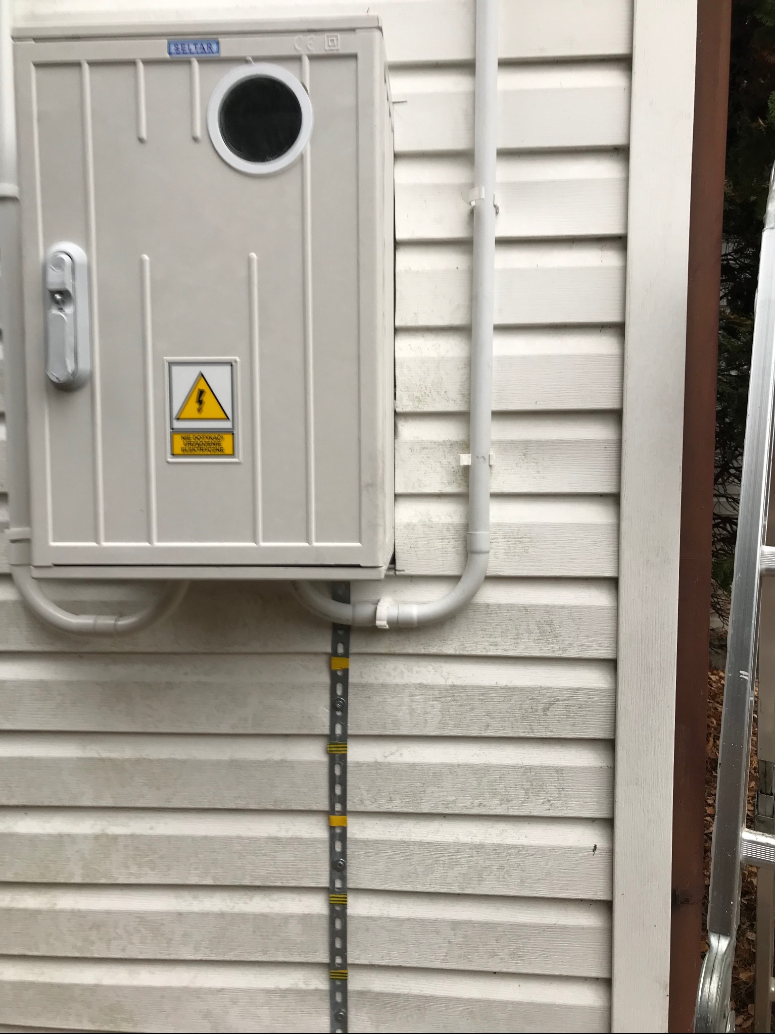 Szara skrzynka elektryczna z okrągłym otworem inspekcyjnym i żółtą naklejką ostrzegawczą, zamontowana na ścianie budynku z sidingu, widoczna rura PCV i drabina.